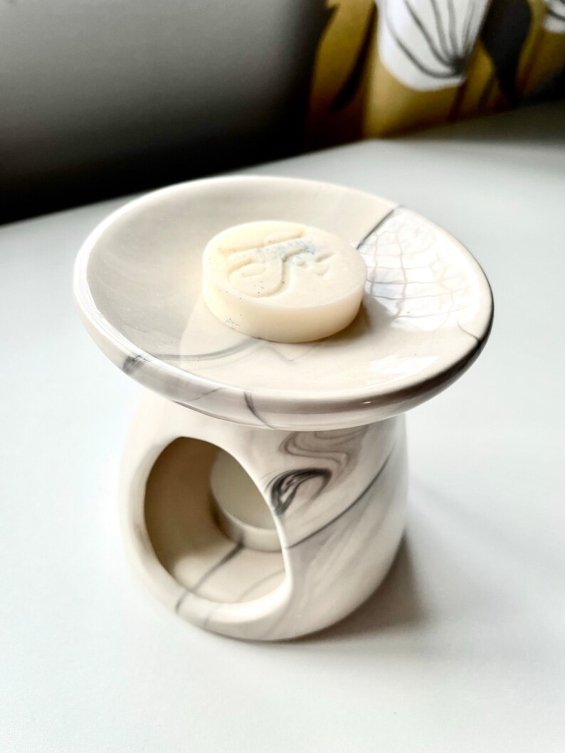 May include: White ceramic wax melt warmer with a marble pattern. The warmer has a round top with a small well for the wax melt and a lower section with a hole for a tea light. A white wax melt is sitting in the well.