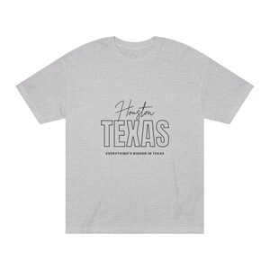 Pode incluir: Uma camiseta cinza mesclada com texto preto que diz "Houston TEXAS" e "Everything's bigger in Texas".