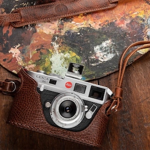 May include: A silver Leica M3 camera with a brown leather case, a brown leather strap, and a black lens cap. The camera is sitting on a wooden surface with a paint palette in the background.