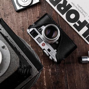 May include: A collection of vintage cameras and photographic equipment on a wooden surface. A Leica M6 camera in a black leather case is the focal point, alongside a light meter and a document with bold text.