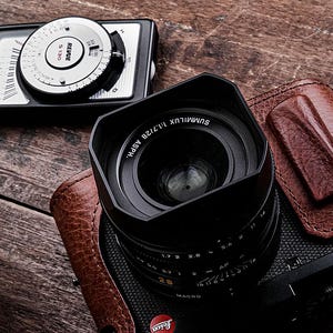 May include: A black camera with a lens and a brown leather case sits on a wooden surface. The lens is labeled SUMMILUX 1:1.7/28 ASPH. and the camera has a red Leica logo. A Revue S 130 light meter is also present.