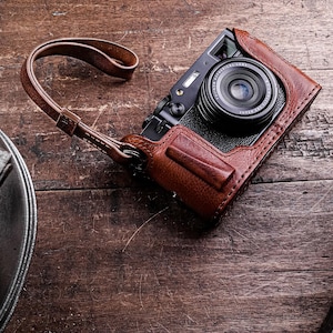 May include: A black digital camera with a brown leather case and strap. The camera lens is centered, and the leather case has a stitched design. The camera is on a wooden surface.