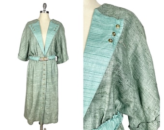 Puede incluir: Un vestido largo de color verde claro con escote en V, mangas cortas y cinturón. El vestido tiene una tela texturizada y está adornado con botones y tachuelas doradas.