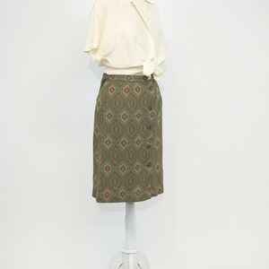 May include: A white button-down shirt with short sleeves tied at the waist over a green and gold geometric patterned skirt.
