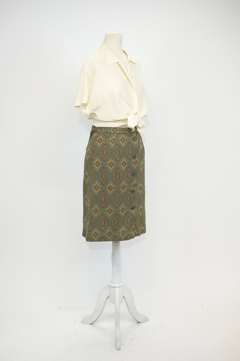 May include: A white button-down shirt with short sleeves tied at the waist over a green and gold geometric patterned skirt.