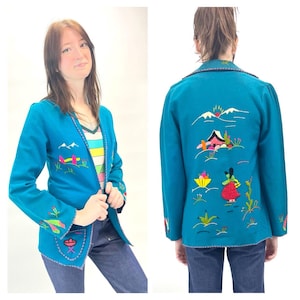 Puede incluir: Una chaqueta azul turquesa con detalles bordados de colores. El bordado representa una escena de paisaje con montañas, una casa y una persona. La chaqueta tiene un bolsillo en el lado izquierdo.