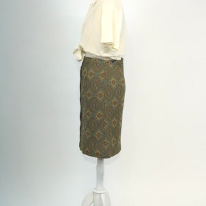 May include: A white blouse with a collar and short sleeves is worn with a green and brown geometric patterned skirt. The skirt is knee-length and has a straight silhouette.