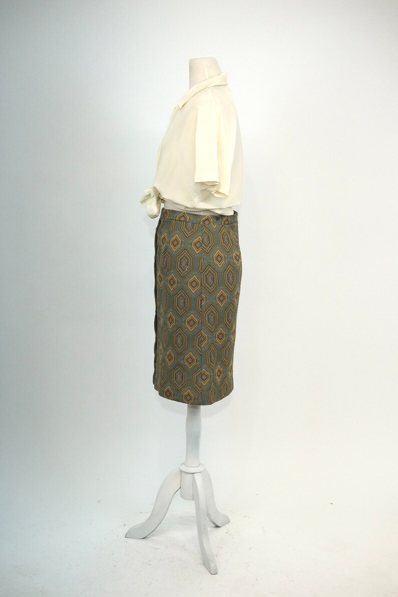 May include: A white blouse with a collar and short sleeves is worn with a green and brown geometric patterned skirt. The skirt is knee-length and has a straight silhouette.