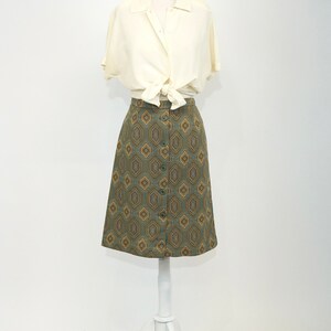 May include: A white button-down shirt tied at the waist over a green and gold geometric patterned skirt.