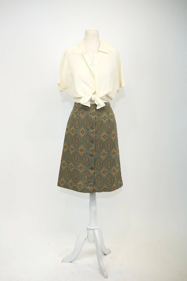 May include: A white button-down shirt tied at the waist over a green and gold geometric patterned skirt.