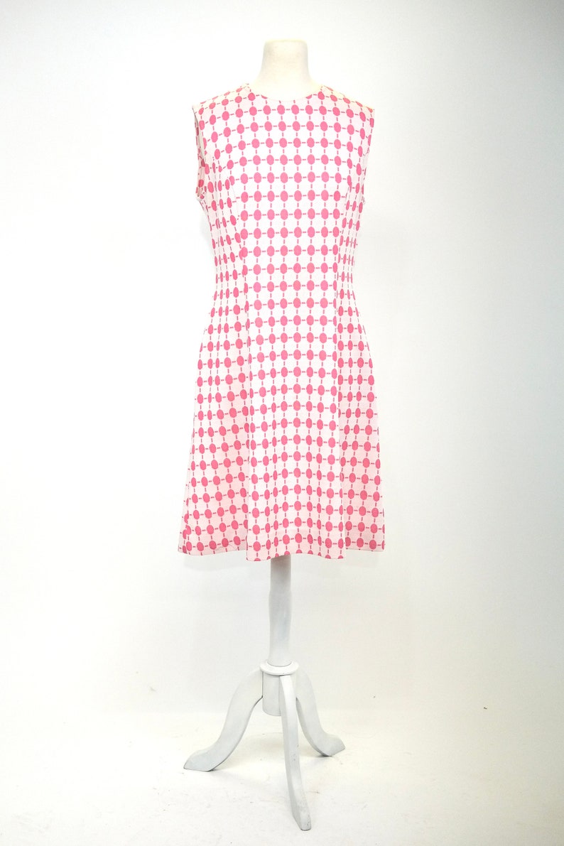 May include: A sleeveless dress with a white background and a pink geometric pattern. The dress has a fitted bodice and a flared skirt.