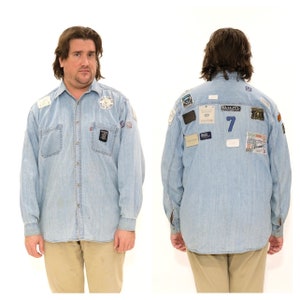 Puede incluir: Camisa vaquera azul claro con múltiples parches y el número 7 en la espalda. La camisa de manga larga tiene dos bolsillos en el pecho y una botonadura frontal. Los parches incluyen logotipos y texto.