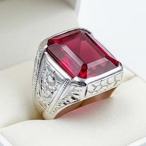 May include: A silver ring with a large, rectangular, red gemstone. The ring is set in a white box. The band features intricate floral and scrollwork designs.