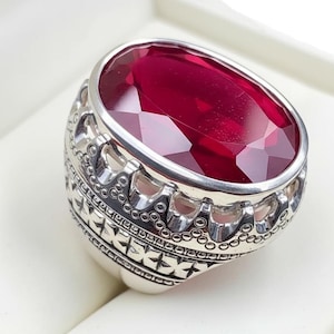 May include: A silver ring with an oval, faceted, deep red gemstone. The ring has an ornate, detailed design around the gemstone and on the band. The ring is set against a white background.