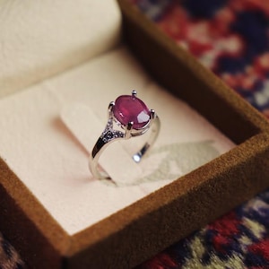 May include: A silver ring with a large, oval-cut ruby gemstone, set in a four-prong setting. The ring is displayed in a brown jewelry box with a cream-colored interior. The background is a patterned rug.