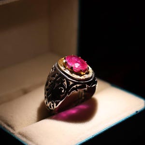 Può includere: Un anello d'argento con una gemma rosa brillante di forma ovale incastonata in una lunetta d'oro. L'anello è esposto in un portagioie blu con interno color crema. L'anello ha un design traforato ornato.