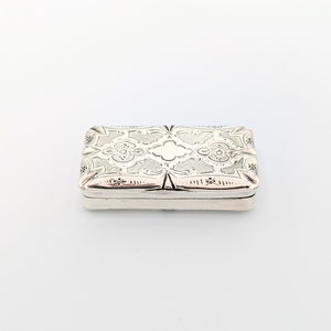 Puede incluir: Una caja rectangular de plata con un diseño grabado intrincado. La caja tiene una tapa con bisagras y está decorada con un patrón floral.