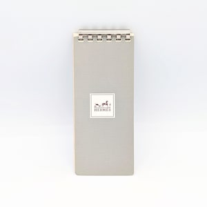 Könnte beinhalten: Ein grauer Notizblock mit einer silbernen Spiralbindung. Der Notizblock hat das Hermes-Logo auf der Vorderseite.