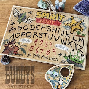 May include: A decorative spirit board with a vintage aesthetic. The board features the alphabet, numbers, and phrases like "Howdy" and "Farewell." The design includes cartoon characters and the text "SPIRIT BOARD."