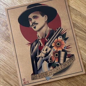 May include: Tattoo-style print featuring a man in a black hat and suit, holding a revolver. A red circle is behind the man's head. The words "IM YOUR HUCKLEBERRY" are on a banner below the image. A flower and a cup are also included.