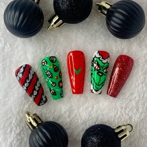 May include: A selection of Christmas-themed false nails with festive designs. The nails feature red, green, and white colours, with patterns including stripes, a Grinch face, and Christmas ornaments. Black Christmas baubles with gold caps surround the nails.
