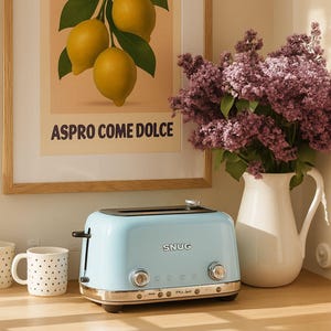 Puede incluir: Una tostadora azul claro SNUG con detalles cromados se encuentra sobre una encimera de madera, junto a una taza blanca con lunares negros. En el fondo, un cuadro enmarcado con limones y un jarrón con flores moradas.