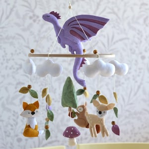 May include: A whimsical baby mobile featuring a purple felt dragon with burgundy wings, white clouds, a fox, a deer, a squirrel, a mushroom, and a green tree. The mobile is suspended from a wooden ring with wooden beads and leaves.