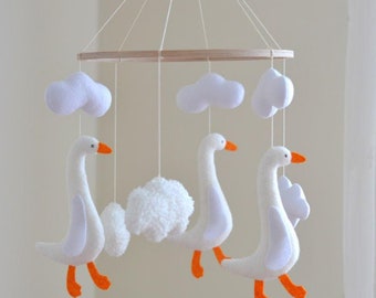 Op de afbeelding: Een witte vilten babymobiel met drie witte ganzen en vier witte wolken die aan een houten ring hangen. De ganzen hebben oranje poten en snavels.