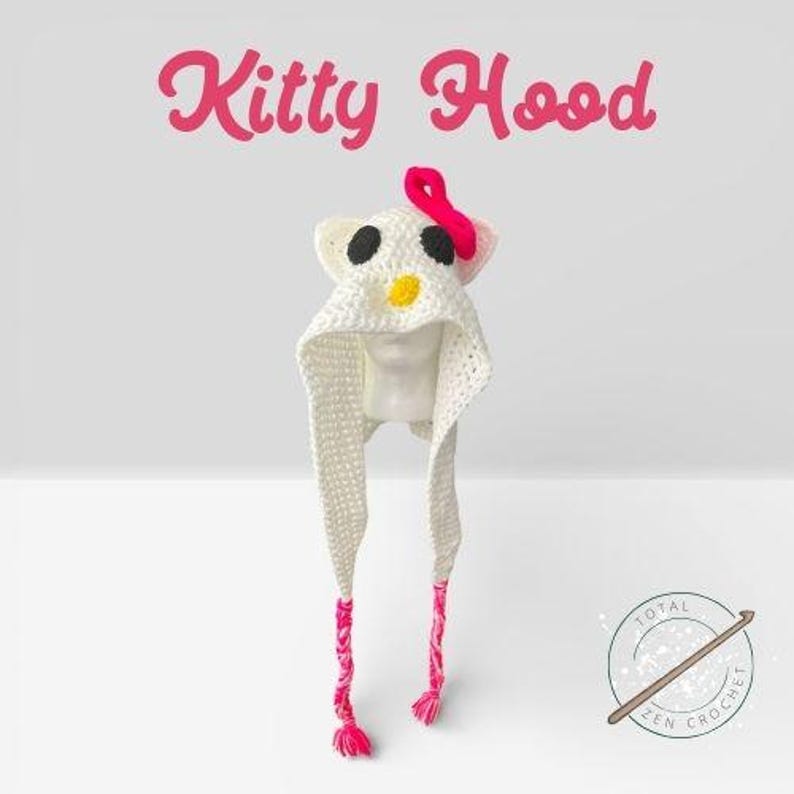 May include: A white crochet Kitty Hood with black eyes, a yellow nose, and a pink bow. The hood has long, braided ties with pink tassels. The words "Kitty Hood" are written in pink script above the hood.