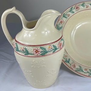 May include: Cream-colored ceramic pitcher and bowl set. The pitcher has a curved handle and a floral design around the middle. The bowl has a matching floral border. Both pieces have embossed floral patterns.