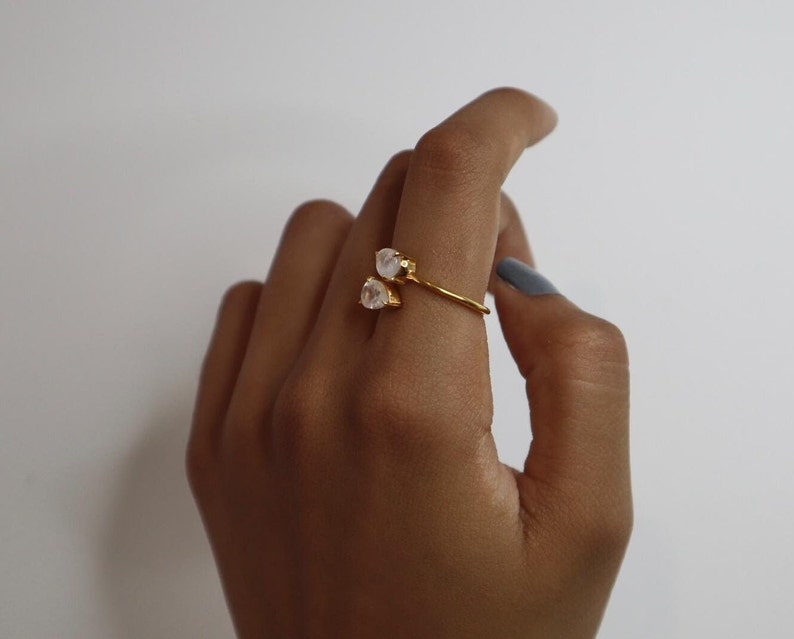 May include: A gold ring with two white stones set on a curved band. The ring is worn on a person's finger.