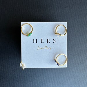 May include: Four gold rings with different coloured gemstones displayed on a white card with the text "HERS Jewellery".