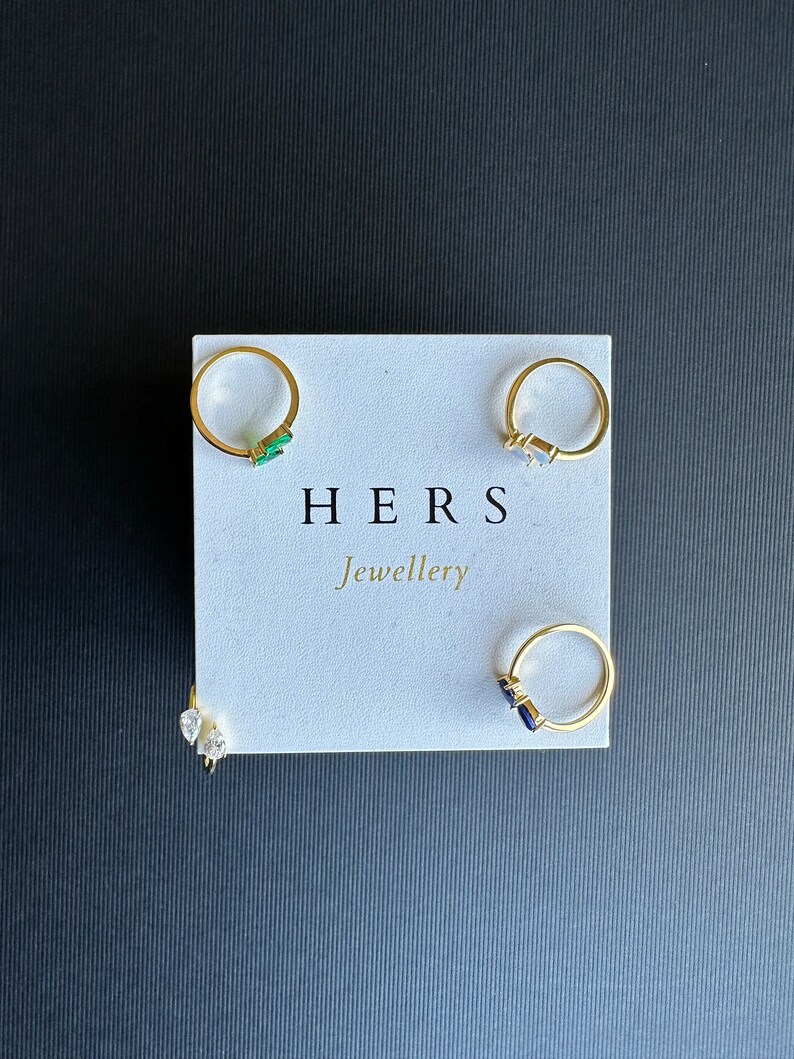 May include: Four gold rings with different coloured gemstones displayed on a white card with the text "HERS Jewellery".