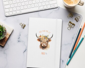 Może przedstawiać: Biały notatnik z ilustracją krowy szkockiej z kwiatową koroną i napisem "YOU HAD ME AT MOO". Notatnik leży na białej marmurowej powierzchni, wraz z klawiaturą, ołówkami i filiżanką kawy. Mierzy około 21 cm długości.