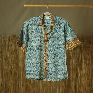 Pode incluir: Uma camisa de manga curta com botões e um padrão azul e branco. A camisa tem uma guarnição castanha e bege na gola e nas mangas. A camisa está pendurada numa barra de madeira.