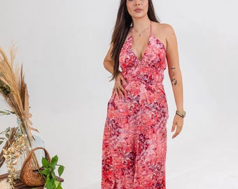 May include: A long, flowing dress with a halter neckline and a vibrant floral pattern in shades of pink and red. The dress has a fitted bodice and a flowing skirt. The model is wearing a gold necklace and bracelet.