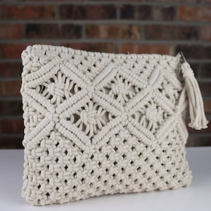 May include: Cream-colored macrame clutch bag with a geometric pattern. The handmade bag features a woven design with a tassel detail. The clutch is made of thick, off-white cotton rope.