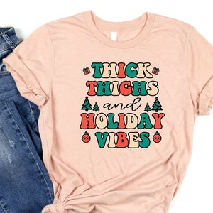 May include: A pink t-shirt with the text "THICK THIGHS and HOLIDAY VIBES" in a retro font with green, red, and blue accents. The shirt is paired with blue jeans.