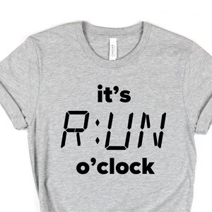 May include: A light gray t-shirt with the text "it's RUN o'clock" in black. The word "RUN" is styled to resemble a digital clock display. The shirt has rolled-up sleeves and a knot at the bottom.