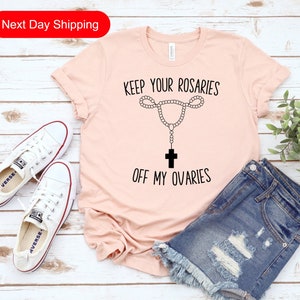 May include: A pink t-shirt with the text "Keep your rosaries off my ovaries" and a graphic of a rosary with a cross hanging from it.