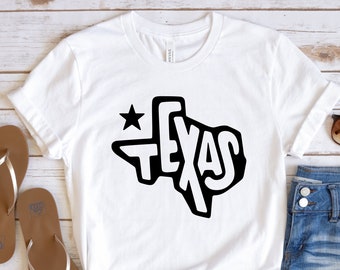 May include: White t-shirt with a black outline of the state of Texas and the word "TEXAS" inside the outline. A small star is above the outline. The shirt is laid out on a wooden surface with a pair of brown flip-flops and denim shorts.