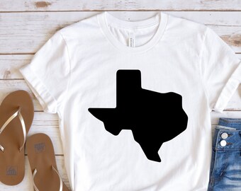 May include: White t-shirt with a black outline of the state of Texas. The shirt is laid out on a white wooden surface with a pair of brown flip-flops, a pair of blue denim shorts, and a pair of brown sunglasses.