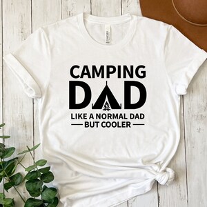 May include: White t-shirt with black text that reads "Camping Dad Like a Normal Dad - But Cooler" with a tent and campfire graphic.