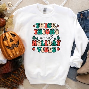 May include: White sweatshirt with the text "THICK THIGHS and HOLIDAY VIBES" in a retro font with colorful accents. The text is surrounded by small Christmas trees and ornaments.