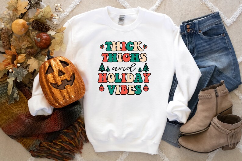 May include: White sweatshirt with the text "THICK THIGHS and HOLIDAY VIBES" in a retro font with colorful accents. The text is surrounded by small Christmas trees and ornaments.