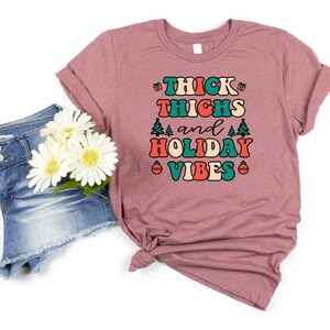 May include: A mauve t-shirt with the text "THICK THIGHS and HOLIDAY VIBES" in a retro font with green, red, and orange accents. The shirt is shown with a pair of blue denim shorts and white flowers.