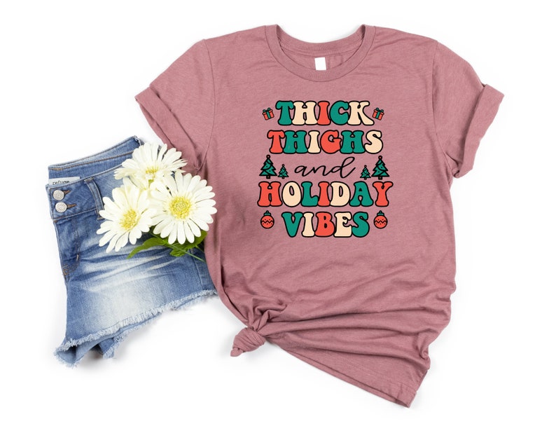May include: A mauve t-shirt with the text "THICK THIGHS and HOLIDAY VIBES" in a retro font with green, red, and orange accents. The shirt is shown with a pair of blue denim shorts and white flowers.