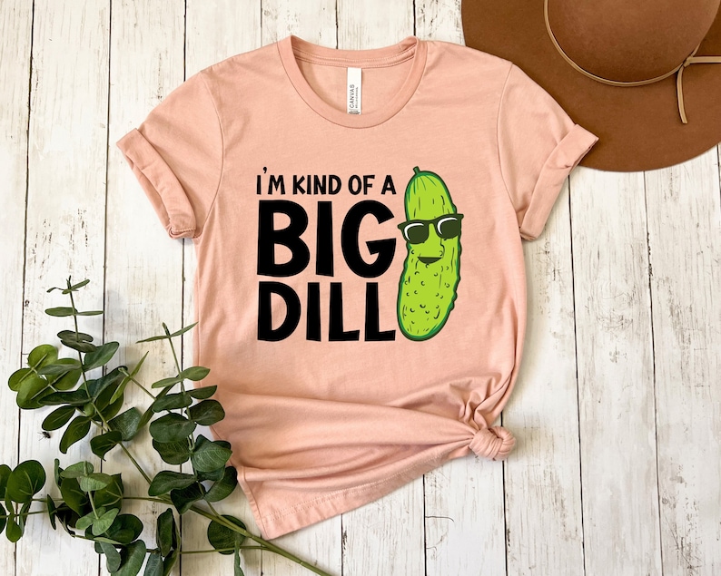 Peut inclure: Un t-shirt rose p&acirc;le avec le texte "I'm kind of a big dill" en noir. Un cornichon de dessin anim&eacute; avec des lunettes de soleil est &agrave; droite du texte.