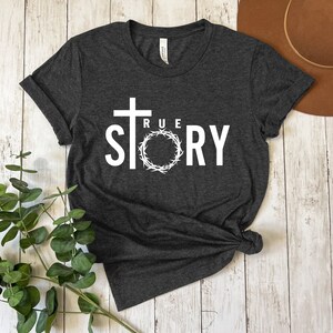 Op de afbeelding: Een donkergrijs T-shirt met een wit grafisch ontwerp. Het ontwerp bevat een kruis, een doornenkroon en de woorden "TRUE STORY".