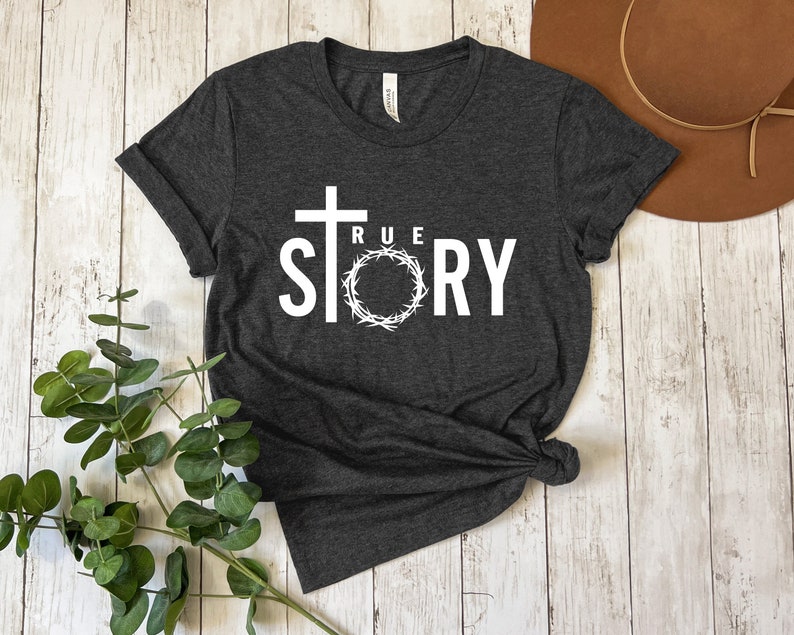 Op de afbeelding: Een donkergrijs T-shirt met een wit grafisch ontwerp. Het ontwerp bevat een kruis, een doornenkroon en de woorden "TRUE STORY".